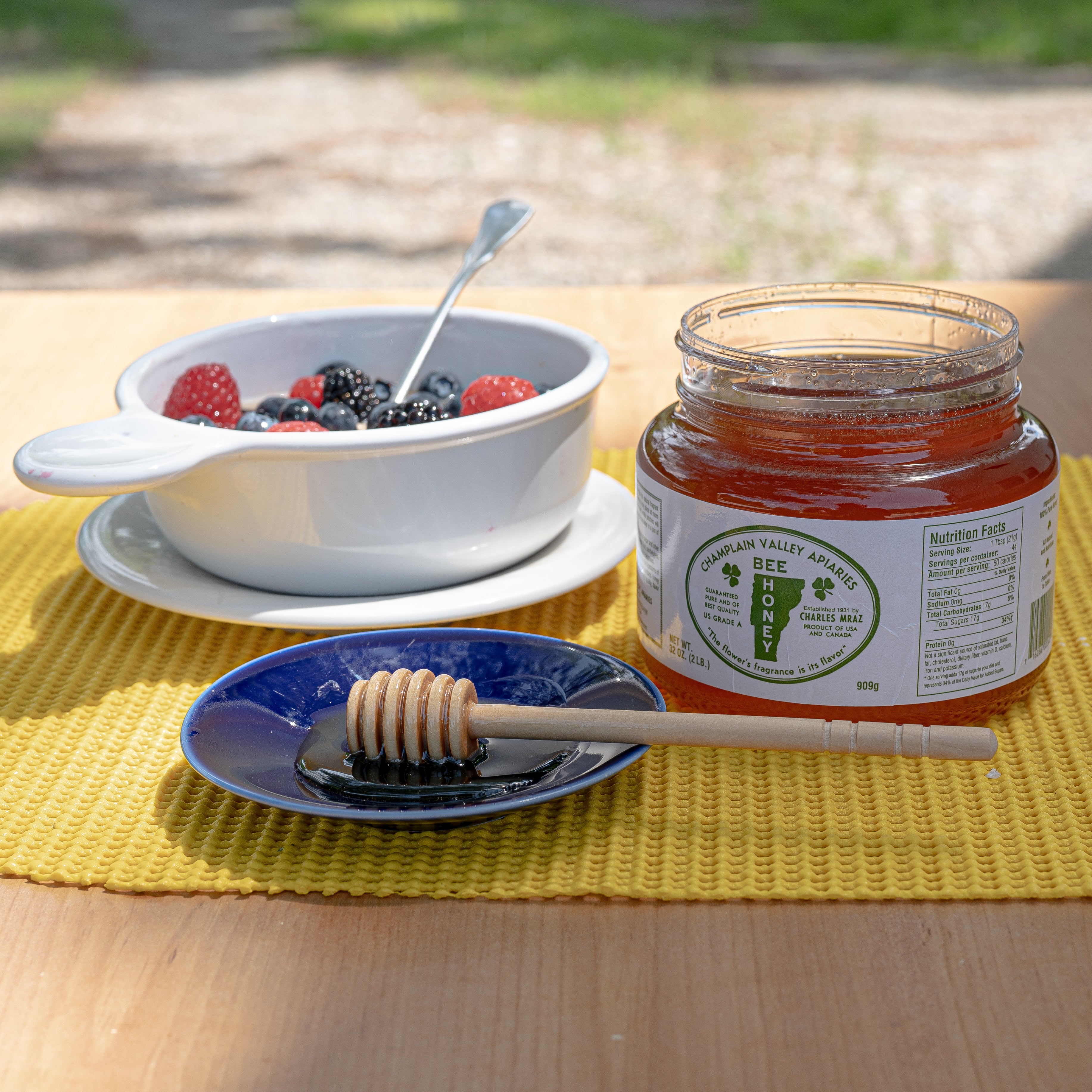 table set outside with a bowl of fruit and a jar of champlain valley apiaries liquid honey and a honey stick on a plate with honey